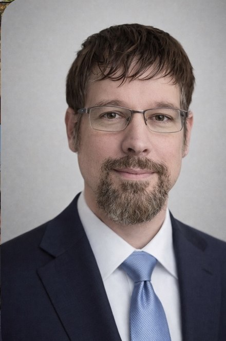 Portrait of author Lance Perry wearing glasses, a dark suit and a blue tie